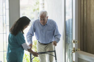A home healthcare worker, a mature woman of Pacific Islander ethnicity, helping a senior man with a walker enter his house through the front door. She is holding the walker, directing him to watch is step.