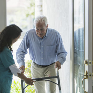 A home healthcare worker, a mature woman of Pacific Islander ethnicity, helping a senior man with a walker enter his house through the front door. She is holding the walker, directing him to watch is step.