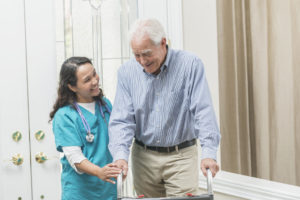 A home healthcare worker helping a senior man with a walker walking through the front door of his house. The nurse is a mature woman, a Pacific Islander, wearing scrubs.