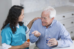 A home healthcare worker helping a senior man in his 70s take his prescription medicine. The patient is in the kitchen, holding a cup of water, smiling at the nurse as he gets ready to swallow the pill in his hand.