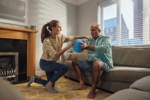 A wife lovingly helps her husband with his strength training and offers encouragement during his physiotherapy in their living room in the North East of England.